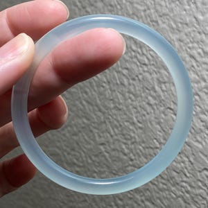 May include: A smooth, translucent, pale blue bangle bracelet with a rounded profile. The circular accessory features a consistent, polished finish throughout its entire circumference, showcasing a clear, icy appearance with subtle light blue tones.