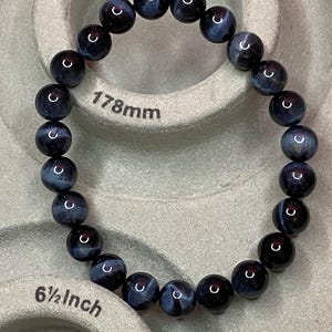 May include: A circular beaded bracelet composed of dark blue and black polished spherical stones. The bracelet is positioned on a measuring tool that displays the length as 178mm and 6 1/2 inches, indicating the size of the jewellery piece.