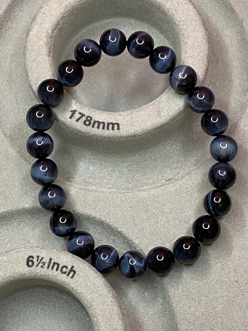 May include: A circular beaded bracelet composed of dark blue and black polished spherical stones. The bracelet is positioned on a measuring tool that displays the length as 178mm and 6 1/2 inches, indicating the size of the jewellery piece.