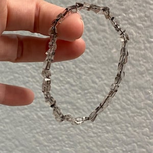 Puede incluir: Una pulsera de cuarzo transparente con un diseño elástico. La pulsera está compuesta por pequeñas cuentas rectangulares translúcidas ensartadas. Las cuentas tienen un aspecto ligeramente ahumado. La pulsera está sujeta por una mano sobre un fondo blanco.