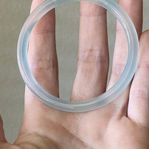 May include: A translucent, pale blue-grey bangle bracelet with a smooth, polished surface. The circular accessory features a rounded profile and a clear, glass-like appearance, reflecting light evenly across its entire circumference.
