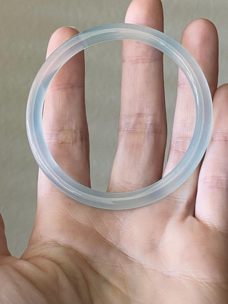 May include: A translucent, pale blue-grey bangle bracelet with a smooth, polished surface. The circular accessory features a rounded profile and a clear, glass-like appearance, reflecting light evenly across its entire circumference.