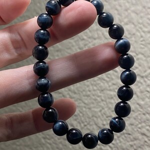 May include: A stretch bracelet composed of round, polished blue tiger eye beads. The beads feature a dark navy blue base colour with subtle, shimmering chatoyant bands of lighter blue and grey that create a reflective, silky appearance throughout.