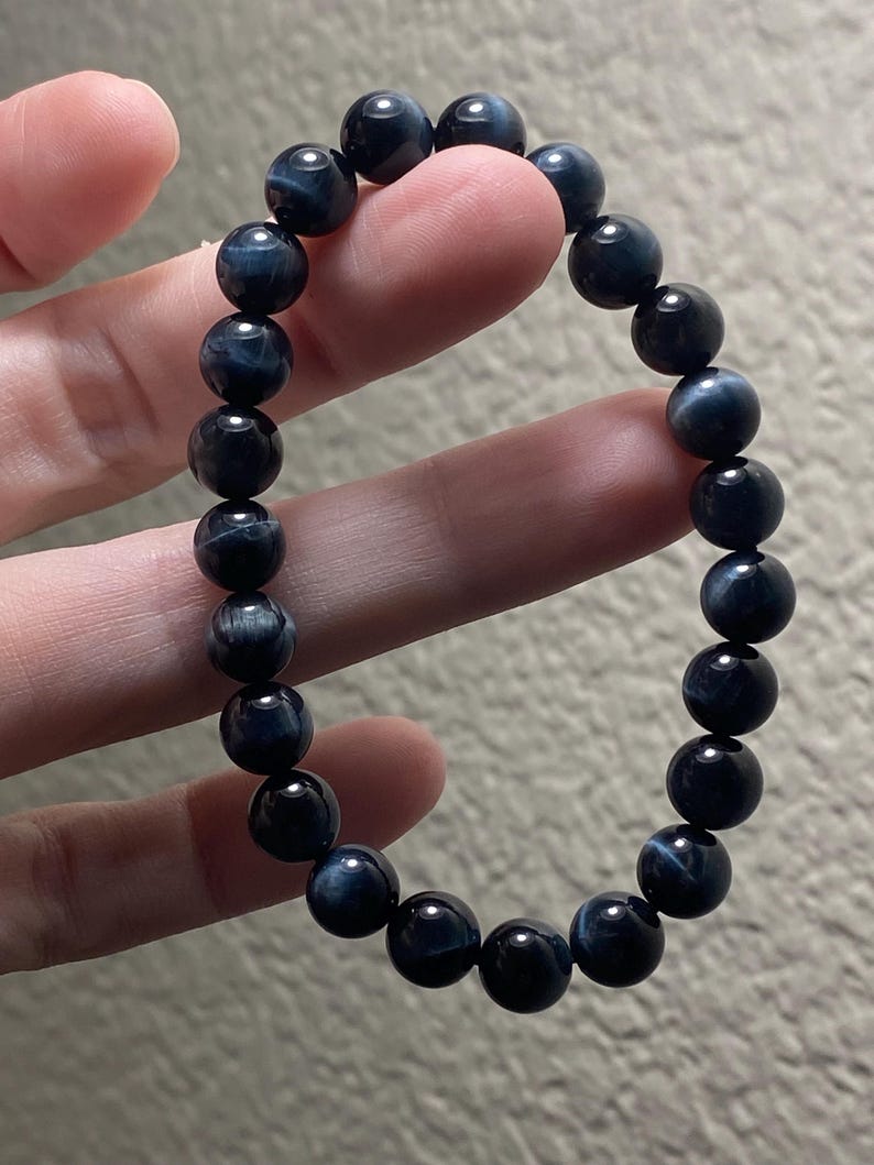 May include: A stretch bracelet composed of round, polished blue tiger eye beads. The beads feature a dark navy blue base colour with subtle, shimmering chatoyant bands of lighter blue and grey that create a reflective, silky appearance throughout.