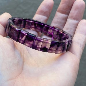 Puede incluir: Una pulsera compuesta por cuentas rectangulares de fluorita translúcida en tonos morados y transparentes. Las cuentas están dispuestas en una banda circular continua, mostrando un zonado de color natural con tonos violetas.