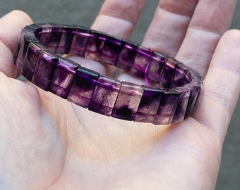 May include: A bracelet composed of rectangular, translucent purple and clear fluorite beads. The beads are arranged in a continuous circular band, showcasing natural color zoning with deep violet hues transitioning into transparent sections.