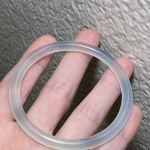May include: A translucent, pale white bangle bracelet with a smooth, rounded surface. The circular accessory features a clear, glass-like appearance with subtle variations in tone, showcasing a simple, continuous band design without any additional ornamentation.