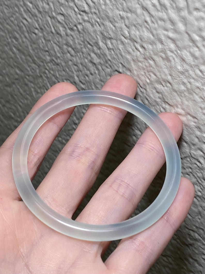 May include: A translucent, pale white bangle bracelet with a smooth, rounded surface. The circular accessory features a clear, glass-like appearance with subtle variations in tone, showcasing a simple, continuous band design without any additional ornamentation.