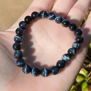 May include: A stretch bracelet composed of round, polished dark blue tiger eye beads. Each bead features a distinct chatoyant band of light reflecting across the surface, creating a shimmering effect. The beads are strung together on an elastic cord.