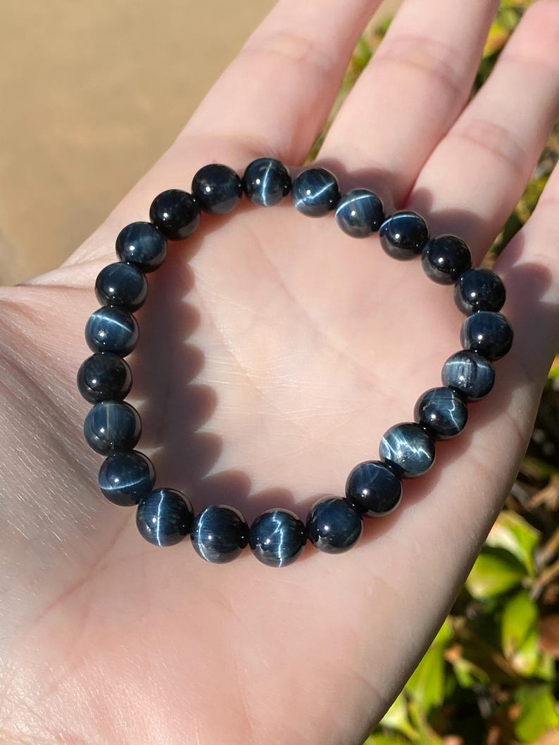 May include: A stretch bracelet composed of round, polished dark blue tiger eye beads. Each bead features a distinct chatoyant band of light reflecting across the surface, creating a shimmering effect. The beads are strung together on an elastic cord.