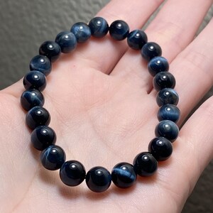 May include: A stretch bracelet composed of round, polished dark blue tiger's eye beads. Each bead displays a chatoyant, shimmering light-reflecting band characteristic of the stone, arranged in a circular pattern on an elastic cord.