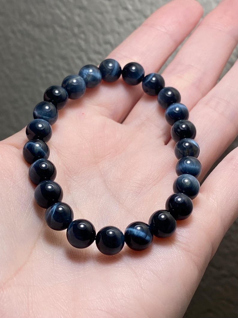 May include: A stretch bracelet composed of round, polished dark blue tiger's eye beads. Each bead displays a chatoyant, shimmering light-reflecting band characteristic of the stone, arranged in a circular pattern on an elastic cord.