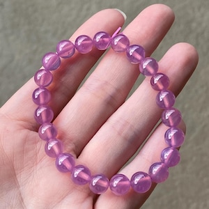 May include: A bracelet made of round, translucent, lavender-colored beads. The beads are strung together to form a complete circle. The bracelet is held in a hand.