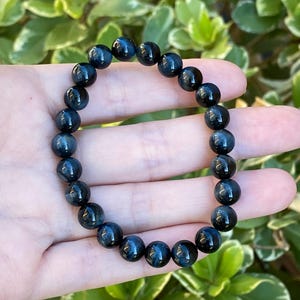 May include: A stretch bracelet composed of round, polished dark blue tiger eye beads. The beads exhibit a chatoyant lustre with deep navy and charcoal tones, arranged in a continuous circular pattern on an elastic cord for a flexible fit.