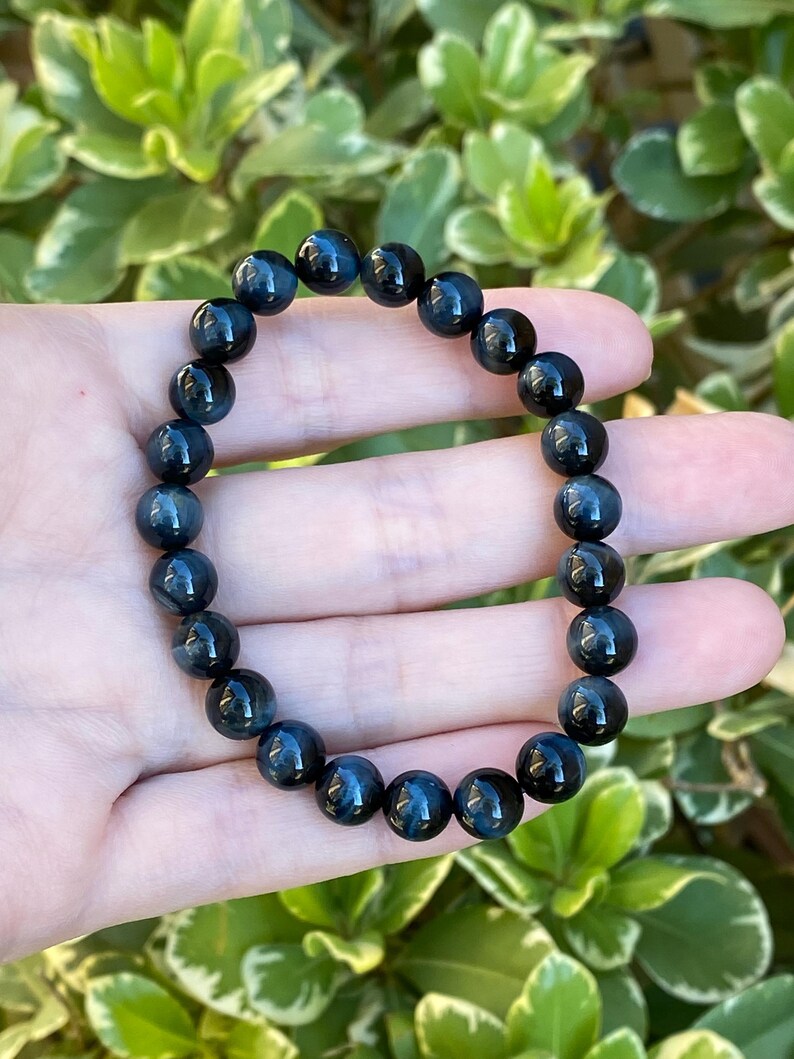 May include: A stretch bracelet composed of round, polished dark blue tiger eye beads. The beads exhibit a chatoyant lustre with deep navy and charcoal tones, arranged in a continuous circular pattern on an elastic cord for a flexible fit.
