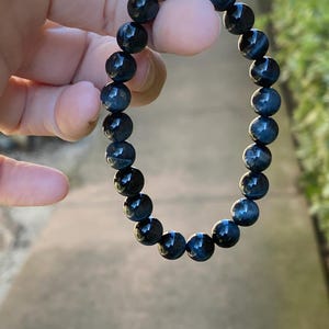 May include: A stretch bracelet composed of round, polished blue tiger eye beads. The beads feature a chatoyant, shimmering effect with deep navy and dark blue tones, arranged in a continuous circular pattern on an elastic cord.