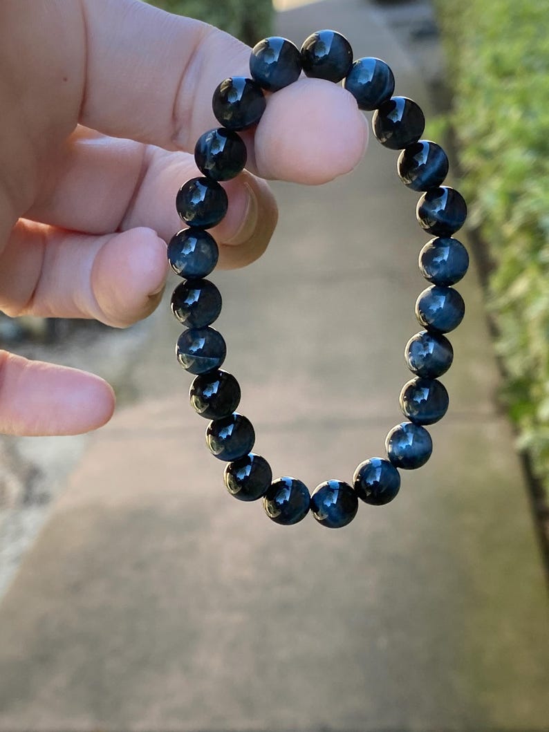 May include: A stretch bracelet composed of round, polished blue tiger eye beads. The beads feature a chatoyant, shimmering effect with deep navy and dark blue tones, arranged in a continuous circular pattern on an elastic cord.