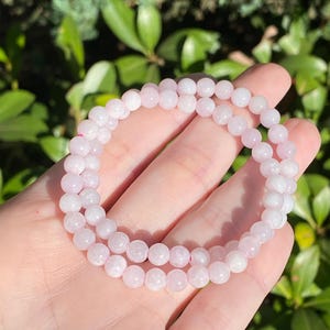 May include: A double-strand bracelet composed of small, smooth, pale pink spherical beads. The beads have a translucent quality and are strung together on a flexible cord, forming two circular loops that rest against the palm of a hand.