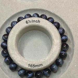 May include: A beaded bracelet made of round, dark blue and black striped gemstone beads. The bracelet is wrapped around a circular beige display tool that features the printed text 6 1/2 Inch and 165mm, indicating the circumference of the jewellery.