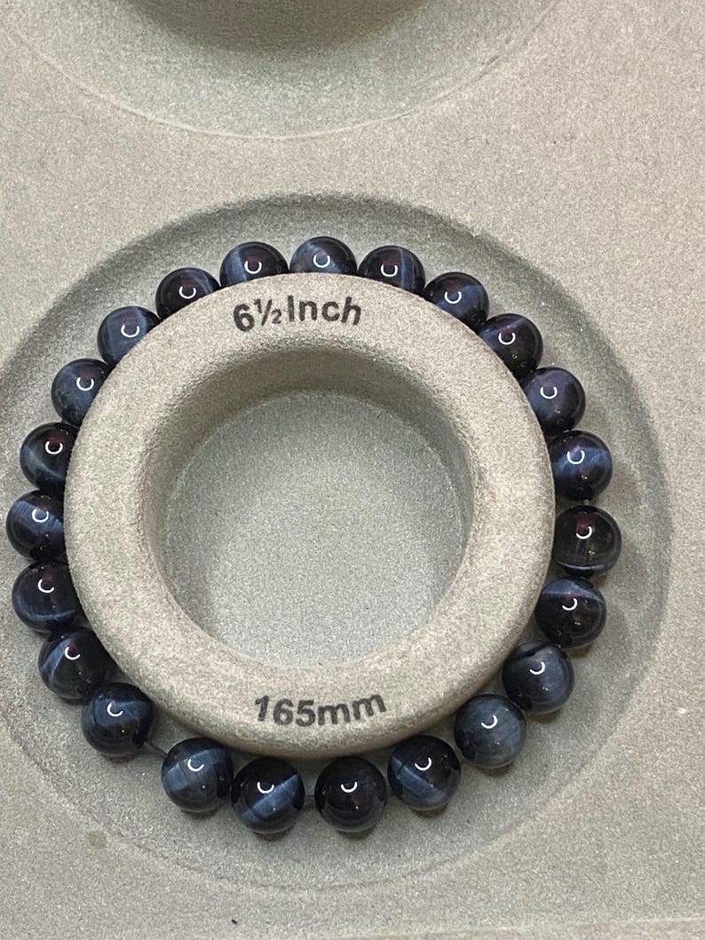 May include: A beaded bracelet made of round, dark blue and black striped gemstone beads. The bracelet is wrapped around a circular beige display tool that features the printed text 6 1/2 Inch and 165mm, indicating the circumference of the jewellery.