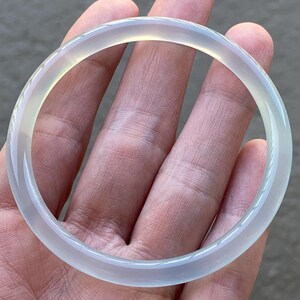 May include: A smooth, circular bangle bracelet crafted from a translucent, pale white stone. The material exhibits a soft, icy appearance with subtle light-gray undertones and a polished, reflective surface that catches the light evenly.