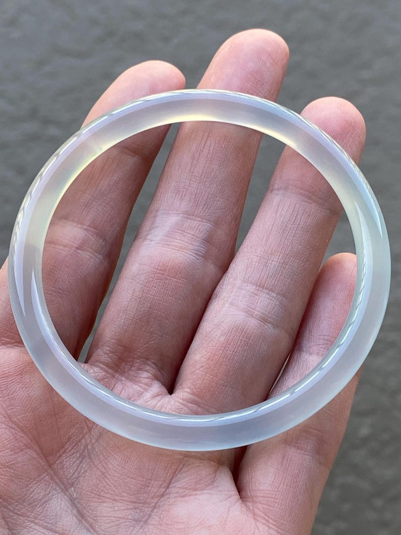May include: A smooth, circular bangle bracelet crafted from a translucent, pale white stone. The material exhibits a soft, icy appearance with subtle light-gray undertones and a polished, reflective surface that catches the light evenly.