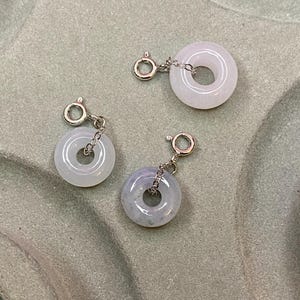 Puede incluir: Tres dijes circulares con anillos de piedra lisa en color lavanda pálido y blanco, suspendidos de anillas y cierres de resorte de metal plateado. Cada pieza consiste en un colgante de piedra pulida en forma de rosquilla con un cierre metálico.