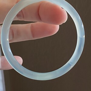 May include: A smooth, circular bangle bracelet with a translucent, pale blue-green appearance. The bracelet features a polished, glossy surface that reflects light, creating a soft glow across its rounded, continuous band structure.