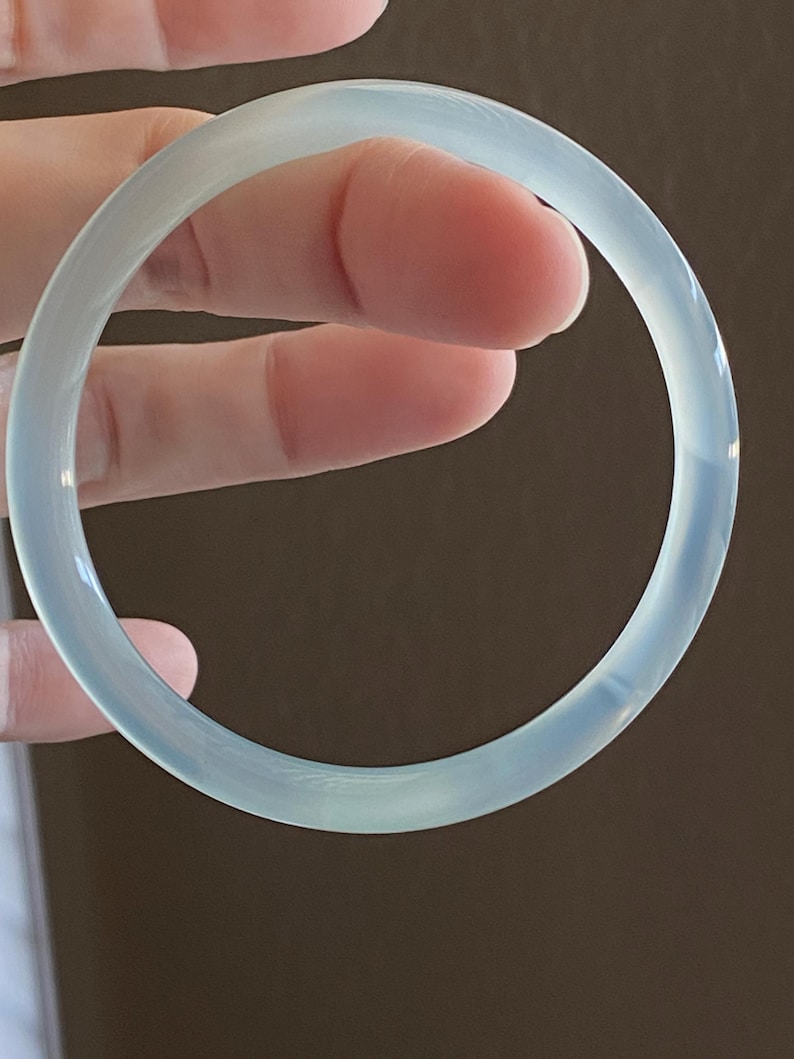 May include: A smooth, circular bangle bracelet with a translucent, pale blue-green appearance. The bracelet features a polished, glossy surface that reflects light, creating a soft glow across its rounded, continuous band structure.