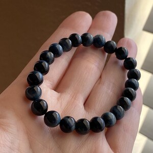 May include: A stretch bracelet composed of round, polished dark blue tiger eye beads. The beads exhibit a smooth, glossy finish with subtle, chatoyant bands of lighter blue and black tones visible throughout the spherical surface of each stone.