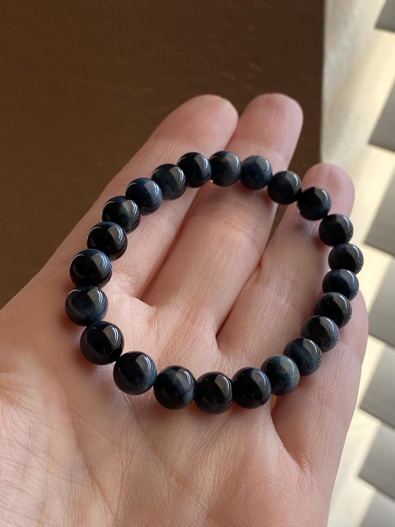May include: A stretch bracelet composed of round, polished dark blue tiger eye beads. The beads exhibit a smooth, glossy finish with subtle, chatoyant bands of lighter blue and black tones visible throughout the spherical surface of each stone.