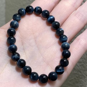 May include: A stretch bracelet composed of round, polished dark blue tiger eye beads. Each bead features a distinct chatoyant shimmer that creates a subtle, light-reflecting band across the surface of the deep blue stone, arranged in a circular loop.
