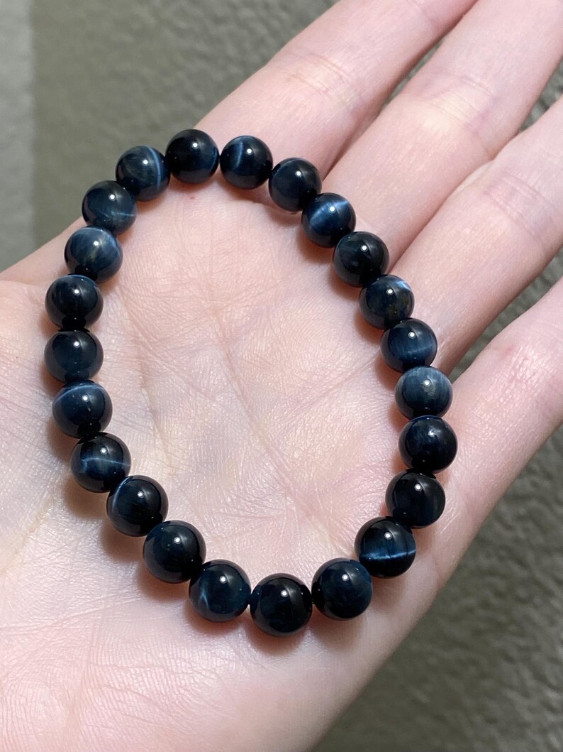 May include: A stretch bracelet composed of round, polished dark blue tiger eye beads. Each bead features a distinct chatoyant shimmer that creates a subtle, light-reflecting band across the surface of the deep blue stone, arranged in a circular loop.
