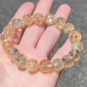 Könnte beinhalten: Ein elastisches Armband aus runden, durchscheinenden, goldgelben Citrinperlen. Jede Perle weist natürliche Einschlüsse sowie eine polierte, reflektierende Oberfläche auf. Die Perlen sind kreisförmig aufgereiht und bilden eine durchgehende Schlaufe aus warmen, honigfarbenen Edelsteinkugeln, die das Sonnenlicht einfangen.