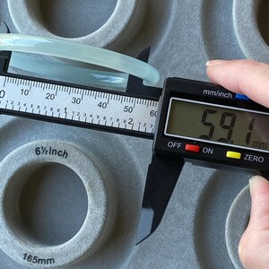 May include: A smooth, pale green bangle bracelet being measured by a black digital caliper. The digital display clearly shows a measurement of 59.1 mm. The bangle is positioned against a grey, circular-patterned surface with a 6.5 inch label.
