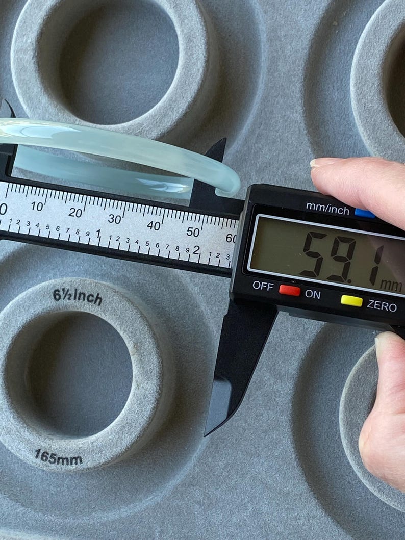 May include: A smooth, pale green bangle bracelet being measured by a black digital caliper. The digital display clearly shows a measurement of 59.1 mm. The bangle is positioned against a grey, circular-patterned surface with a 6.5 inch label.