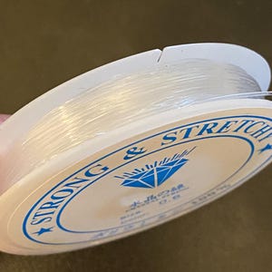 Op de afbeelding: Een spoel met transparante, elastische draad voor kralen. De witte spoel heeft blauwe tekst met de tekst "STRONG & STRETCH" en Japanse tekst. De draad is strak opgerold en lijkt transparant.
