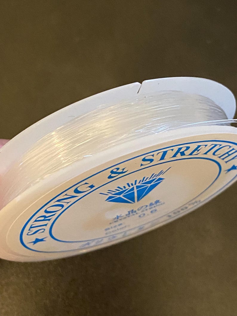 Op de afbeelding: Een spoel met transparante, elastische draad voor kralen. De witte spoel heeft blauwe tekst met de tekst "STRONG & STRETCH" en Japanse tekst. De draad is strak opgerold en lijkt transparant.