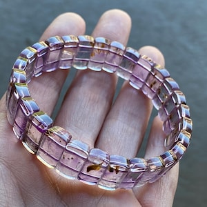 Puede incluir: Una pulsera de amatista translúcida con un diseño rectangular. La pulsera está compuesta por cuentas rectangulares individuales, de color púrpura claro, con detalles dorados. Las cuentas tienen inclusiones y están unidas para formar una forma circular.