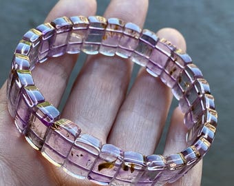 May include: A translucent amethyst bracelet with a rectangular design. The bracelet is composed of individual, light purple, rectangular beads with golden accents. The beads have inclusions and are strung together to form a circular shape.