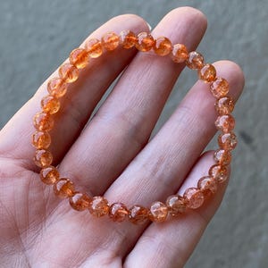 May include: A stretch bracelet composed of small, round, translucent orange sunstone beads. Each bead features internal shimmering inclusions that catch the light, creating a sparkling effect throughout the circular arrangement of the gemstone jewellery.