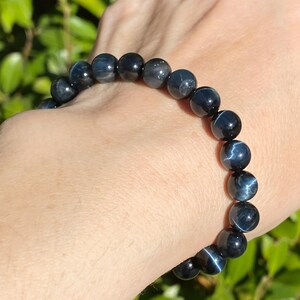 May include: A stretch bracelet composed of round, polished blue tiger's eye beads. Each bead features a distinct chatoyant lustre, displaying shifting bands of deep navy and slate blue tones that create a reflective, cat's eye effect across the surface.