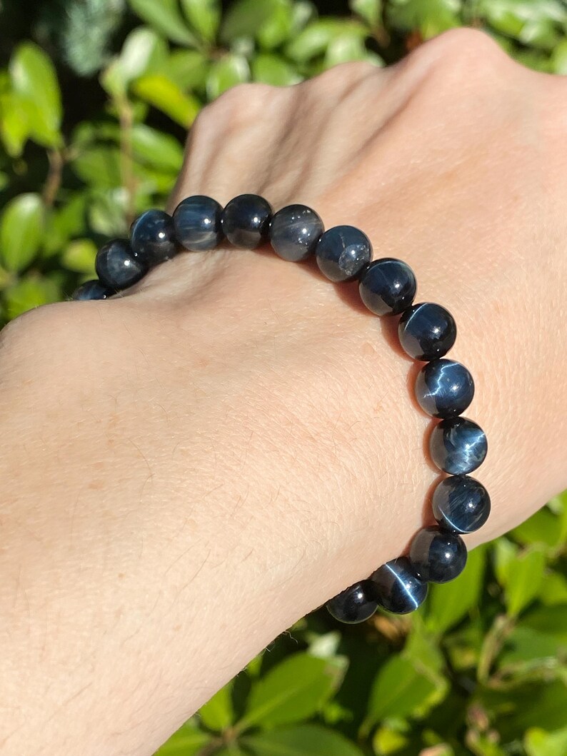 May include: A stretch bracelet composed of round, polished blue tiger's eye beads. Each bead features a distinct chatoyant lustre, displaying shifting bands of deep navy and slate blue tones that create a reflective, cat's eye effect across the surface.