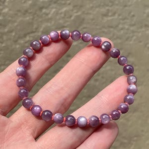 May include: A stretch bracelet composed of small, round, polished purple beads with a subtle chatoyant sheen. The beads are strung on a thin, bright orange elastic cord that is visible between each individual bead in the circular arrangement.
