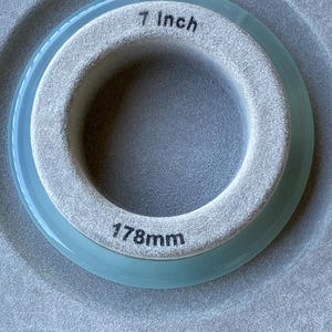 May include: A circular ring with a light blue translucent outer edge and a textured grey inner band. The inner band features the printed text 7 Inch at the top and 178mm at the bottom, indicating the diameter of the circular object.