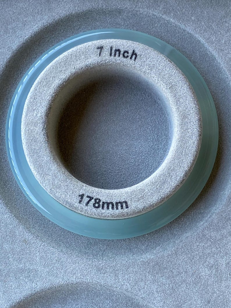 May include: A circular ring with a light blue translucent outer edge and a textured grey inner band. The inner band features the printed text 7 Inch at the top and 178mm at the bottom, indicating the diameter of the circular object.