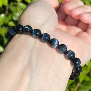 May include: A stretch bracelet composed of round, polished blue tiger's eye beads. Each bead features a dark navy blue base colour with a distinct, shimmering chatoyant band that reflects light, creating a subtle, silky, and linear pattern.
