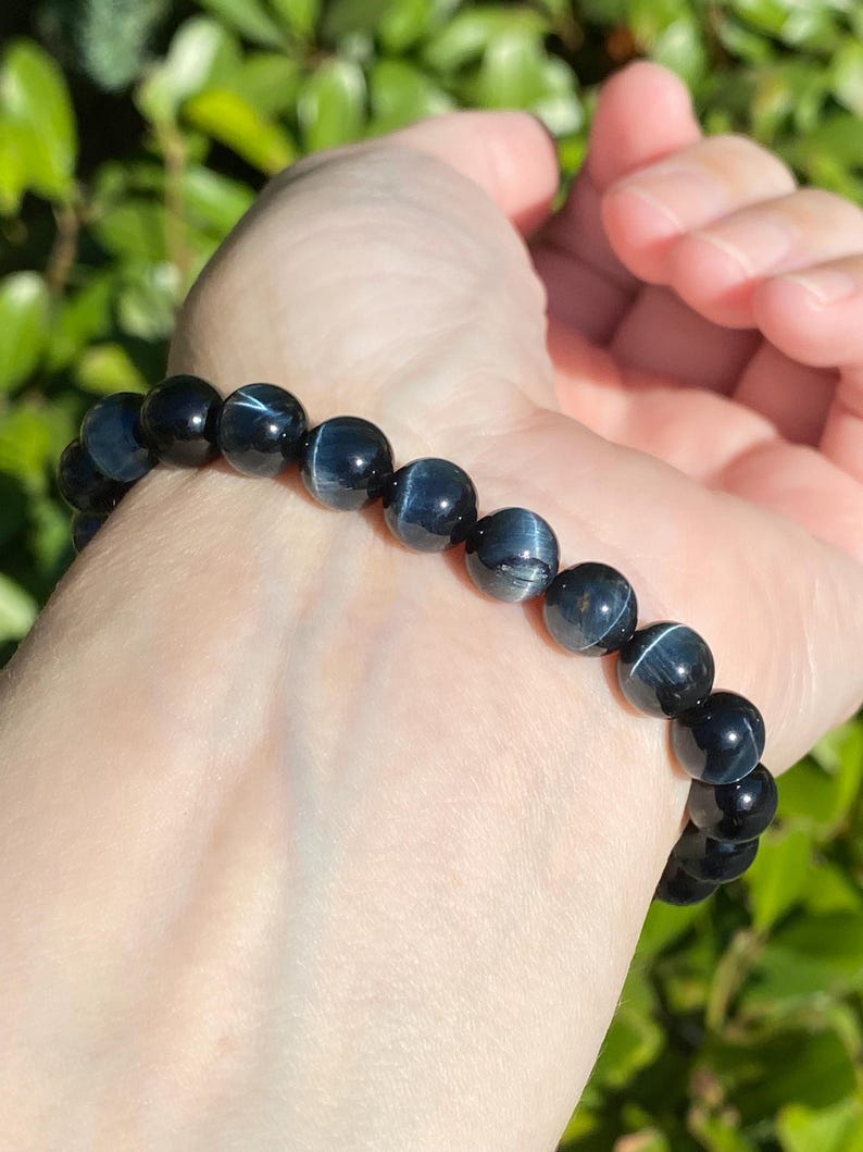 May include: A stretch bracelet composed of round, polished blue tiger's eye beads. Each bead features a dark navy blue base colour with a distinct, shimmering chatoyant band that reflects light, creating a subtle, silky, and linear pattern.