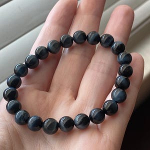 May include: A stretch bracelet composed of round, polished blue tiger's eye beads. The beads feature a chatoyant, silky lustre with deep navy and slate blue tones, arranged in a continuous circular pattern on an elastic cord for a flexible fit.