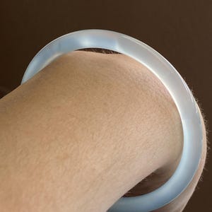 May include: A smooth, translucent, pale blue bangle bracelet with a rounded profile. The material appears semi-transparent, allowing light to pass through the surface, creating a soft glow and a consistent, polished finish around the entire circular band.